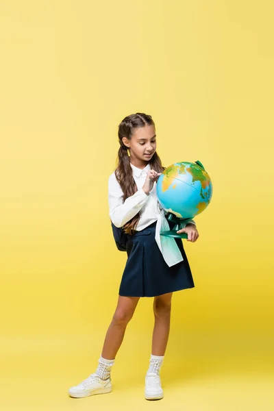 Colegiala positiva con mochila mirando globo sobre fondo amarillo - foto de stock