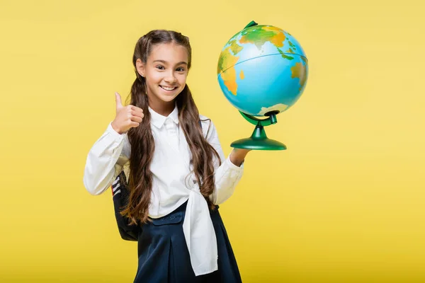Smiling schoolgirl showing like and holding globe isolated on yellow — Stock Photo