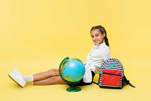 Lächelndes Schulmädchen sitzt neben Globus und Rucksack auf gelbem Hintergrund — Stockfoto