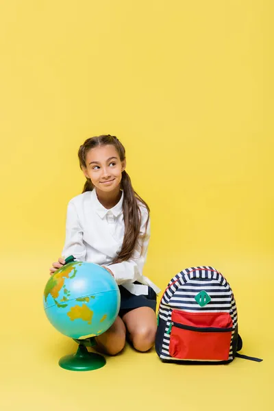 Smiling schoolkid sitting near globe and backpack on yellow background — Stock Photo