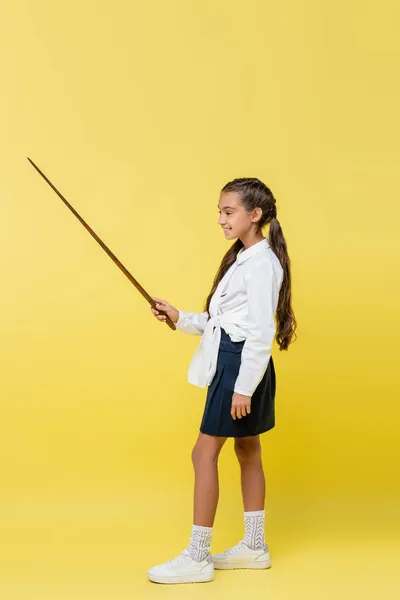 Full length of schoolchild in skirt holding pointer on yellow background — Stock Photo
