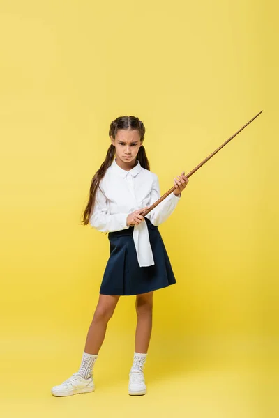 Écolière sérieuse en jupe tenant pointeur sur fond jaune — Photo de stock