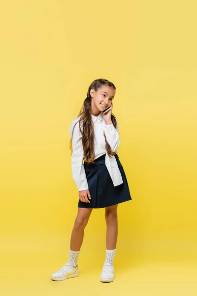 Улыбающаяся школьница в рубашке и юбке разговаривает по мобильному телефону на желтом фоне — стоковое фото