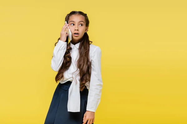 Astonished schoolgirl talking on mobile phone and looking away on yellow background — Stock Photo