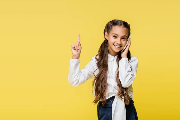 Smiling schoolkid talking on smartphone and pointing with finger isolated on yellow — Stock Photo