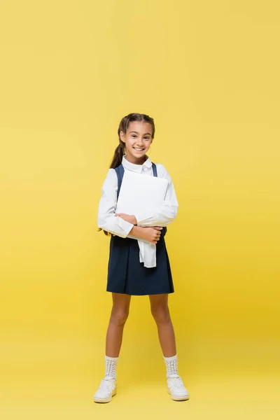 Full length of smiling schoolgirl with backpack holding laptop on yellow background — Stock Photo