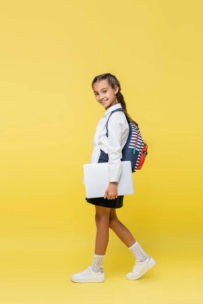 Preadolescente colegiala con mochila y portátil caminando sobre fondo amarillo - foto de stock