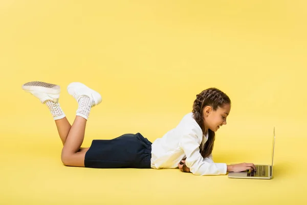 Vista lateral de un niño en edad escolar positiva usando un ordenador portátil mientras está acostado sobre un fondo amarillo - foto de stock