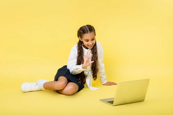 Улыбающаяся школьница получает видеозвонок на ноутбуке на желтом фоне — стоковое фото