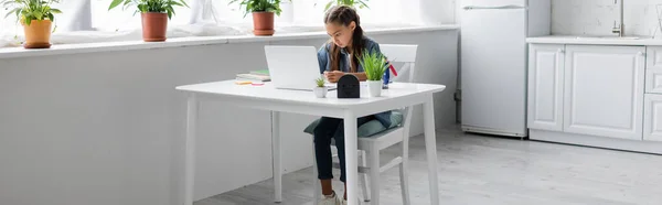 Niño usando el ordenador portátil mientras hace el trabajo escolar en la cocina, pancarta - foto de stock