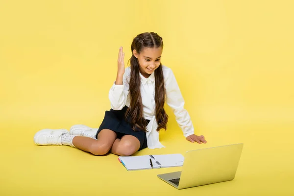 Lächelndes Schulkind hebt während Videoanruf auf Laptop neben Notizbuch auf gelbem Hintergrund die Hand — Stockfoto