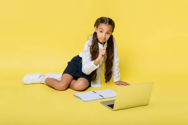 Pensive schoolkid hält Stift in der Nähe von Laptop und kopieren Buch auf gelbem Hintergrund — Stockfoto