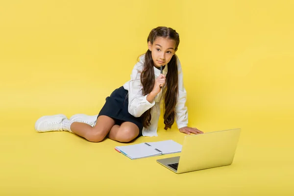 Pensive schoolkid holding pen near laptop and copy book on yellow background — Stock Photo