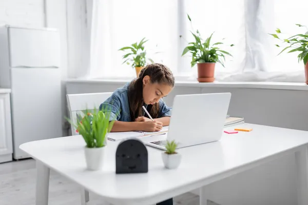 Schulkind schreibt auf Notizbuch neben Laptop und Klebezettel auf Tisch in Küche — Stockfoto
