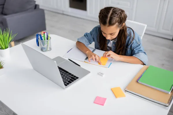 Mädchen klebt Zettel auf Notizbuch in der Nähe von Schreibwaren und Laptop zu Hause — Stockfoto