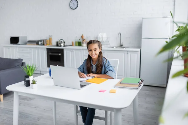 Ребенок-подросток улыбается в камеру рядом с ноутбуками и ноутбуком на кухне — стоковое фото
