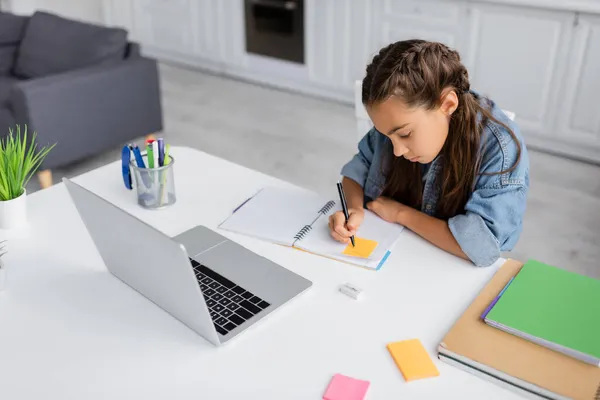 Niño preadolescente escribiendo en nota adhesiva en el portátil cerca de la computadora portátil en casa - foto de stock