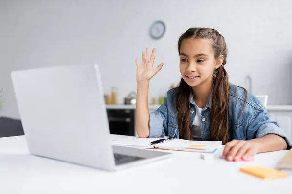 Bambino sorridente che fa videochiamata su computer portatile vicino a notebook durante la scuola di casa in cucina — Foto stock