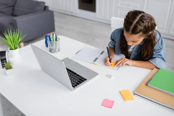 Preteen schoolkid Schreiben auf Notizbuch mit Haftnotiz in der Nähe von Laptop und Pflanzen auf dem Tisch zu Hause — Stockfoto