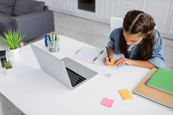 Preadolescente escolar escribir en el cuaderno con nota adhesiva cerca de la computadora portátil y plantas en la mesa en casa - foto de stock