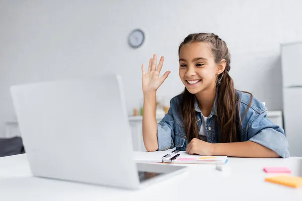 Positive kid waving hand during video chat on laptop near blurred sticky notes at home — Stock Photo
