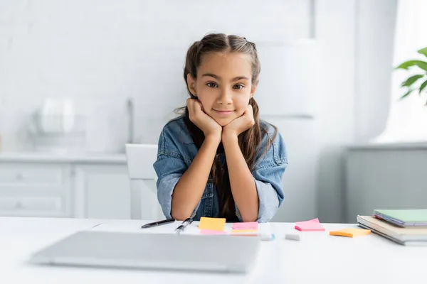 Preteen schoolkid looking at camera near blurred laptop and notebooks in kitchen — Stock Photo