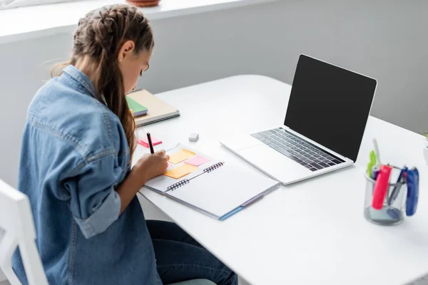 Escolar desfocado escrevendo no notebook com notas pegajosas perto do laptop com tela em branco em casa — Fotografia de Stock