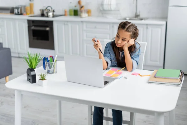 Bored schoolkid holding pen near notebooks and laptop in kitchen — Stock Photo