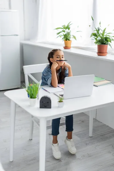 Preadolescente niño sosteniendo pluma cerca de los labios mientras mira la cámara cerca de la computadora portátil y portátiles en casa - foto de stock