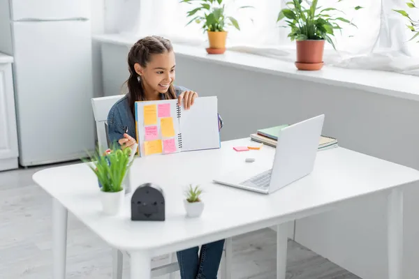 Lächelndes Schulmädchen hält Notizbuch mit klebrigen Notizen in der Nähe des Laptops während des Online-Unterrichts zu Hause — Stockfoto