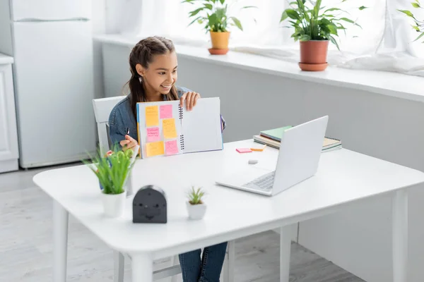 Smiling schoolgirl holding notebook with sticky notes near laptop during online education at home — Stock Photo