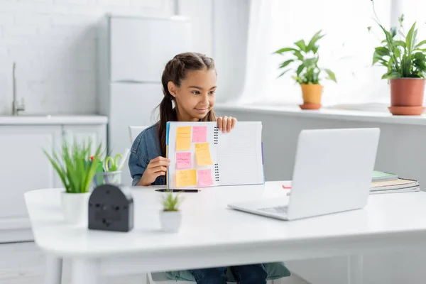Smiling kid showing notebook with sticky notes at laptop during video call at home — Stock Photo