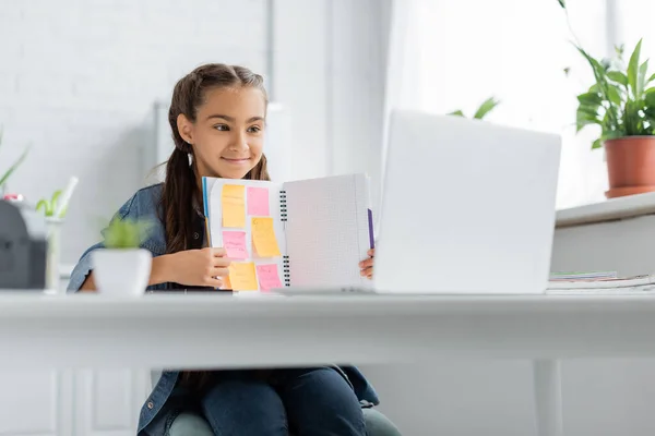 Niño de la escuela sosteniendo el cuaderno con notas adhesivas cerca de la computadora portátil borrosa durante la videollamada en casa - foto de stock