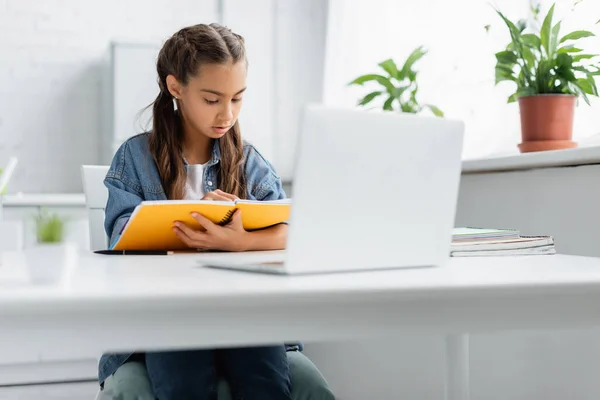 Chica preadolescente sosteniendo portátil cerca borrosa portátil durante la educación en línea en el hogar - foto de stock
