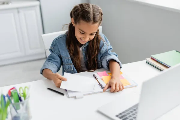 Felice bambino strappando carta vicino notebook con appunti appiccicosi e laptop sfocato a casa — Foto stock