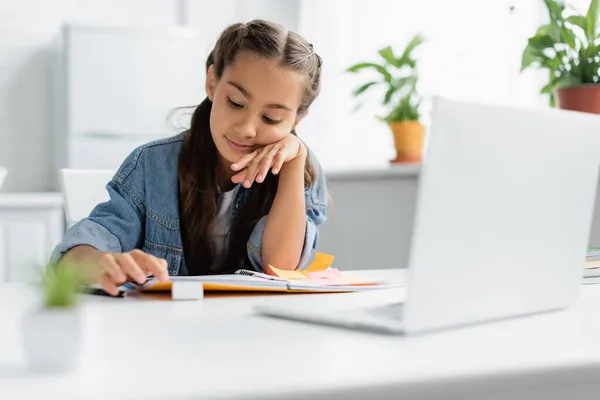 Kind schaut bei Hausaufgaben in der Küche neben Laptop auf Kopierbuch — Stockfoto