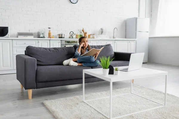 Thoughtful kid holding notebook while sitting on couch near laptop on coffee table at home — Stock Photo