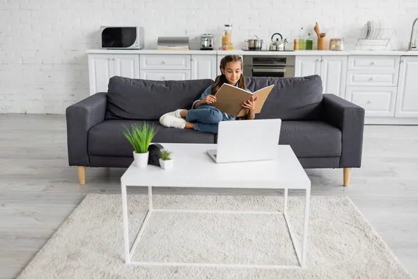 Preteen Kind hält Notizbuch auf Couch neben Laptop auf Couchtisch in Küche — Stockfoto