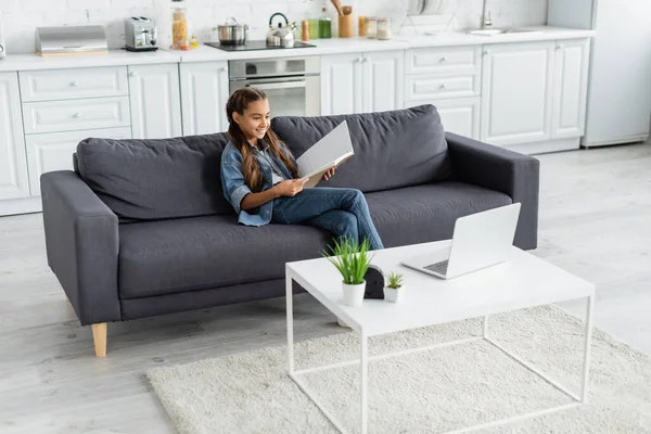 Smiling preteen child holding notebook near laptop on coffee table at home — Stock Photo