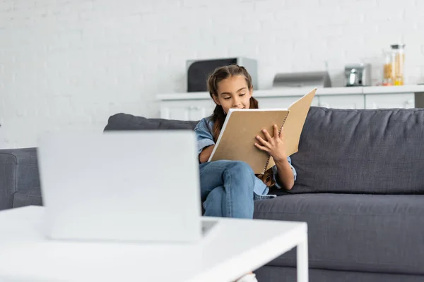 Schoolgirl holding notebook near blurred laptop at home — Stock Photo