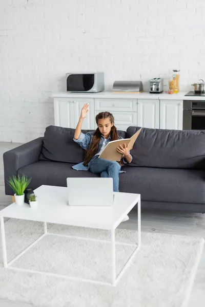 Niño preadolescente con portátil levantando la mano cerca de la computadora portátil en el sofá en casa - foto de stock