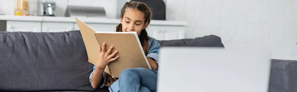 Bambino che tiene il taccuino vicino al computer portatile offuscato a casa, banner — Foto stock