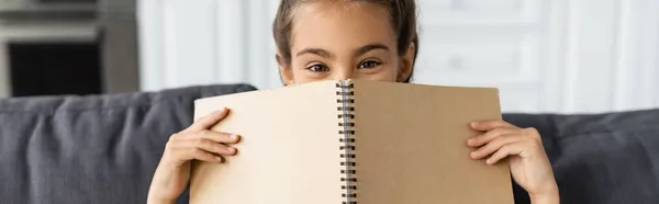 Preteen kid covering face with notebook on couch at home, banner — Stock Photo