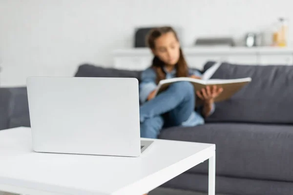 Laptop on coffee table near blurred child with notebook on couch — Stock Photo