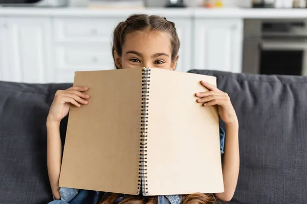 Preteen kid cover face with notebook and looking at couch at home — Stockfoto