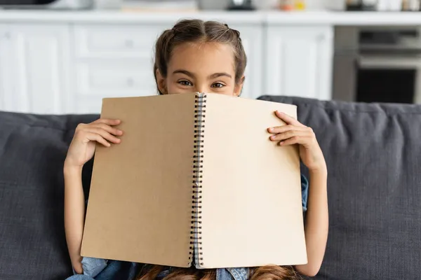 Preadolescente niño cubriendo la cara con portátil y mirando a la cámara en el sofá en casa - foto de stock