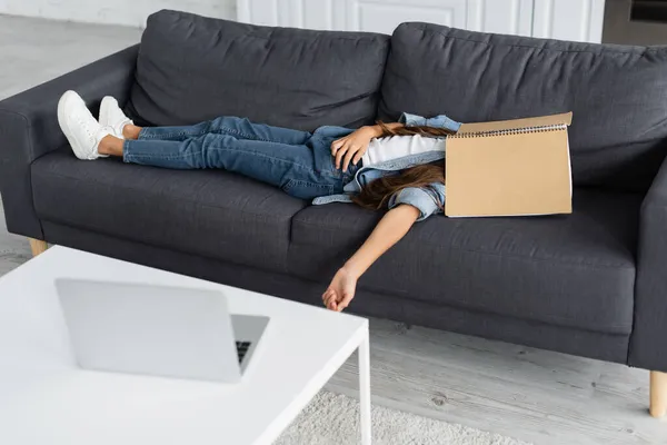 Mädchen mit Notizbuch im Gesicht liegt auf Couch neben verschwommenem Laptop zu Hause — Stockfoto