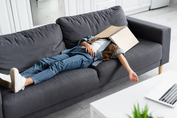 Preteen kid with notebook on face lying on couch near laptop on coffee table at home — Stock Photo
