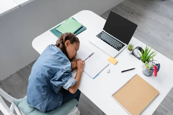 Вид сверху ребенка, спящего рядом с ноутбуком и тетрадями на столе дома — стоковое фото