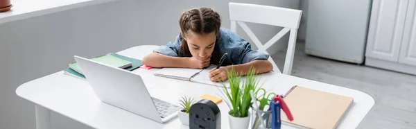 Ребенок пишет на ноутбуке возле ноутбука и канцелярских принадлежностей дома, баннер — стоковое фото